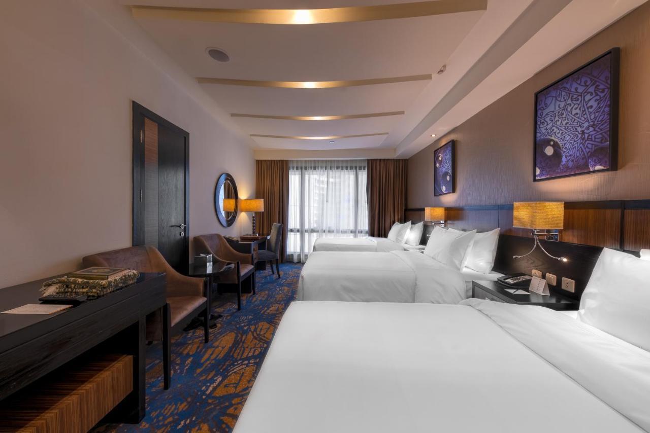 Al Ghufran Safwah Hotel Makkah Meca Exterior foto The photo shows a hotel room featuring two large beds with white bedding. There are two bedside tables with lamps beside each bed. On one side of the room, there is a comfortable chair and a small table, while a work desk can also be seen. The floori